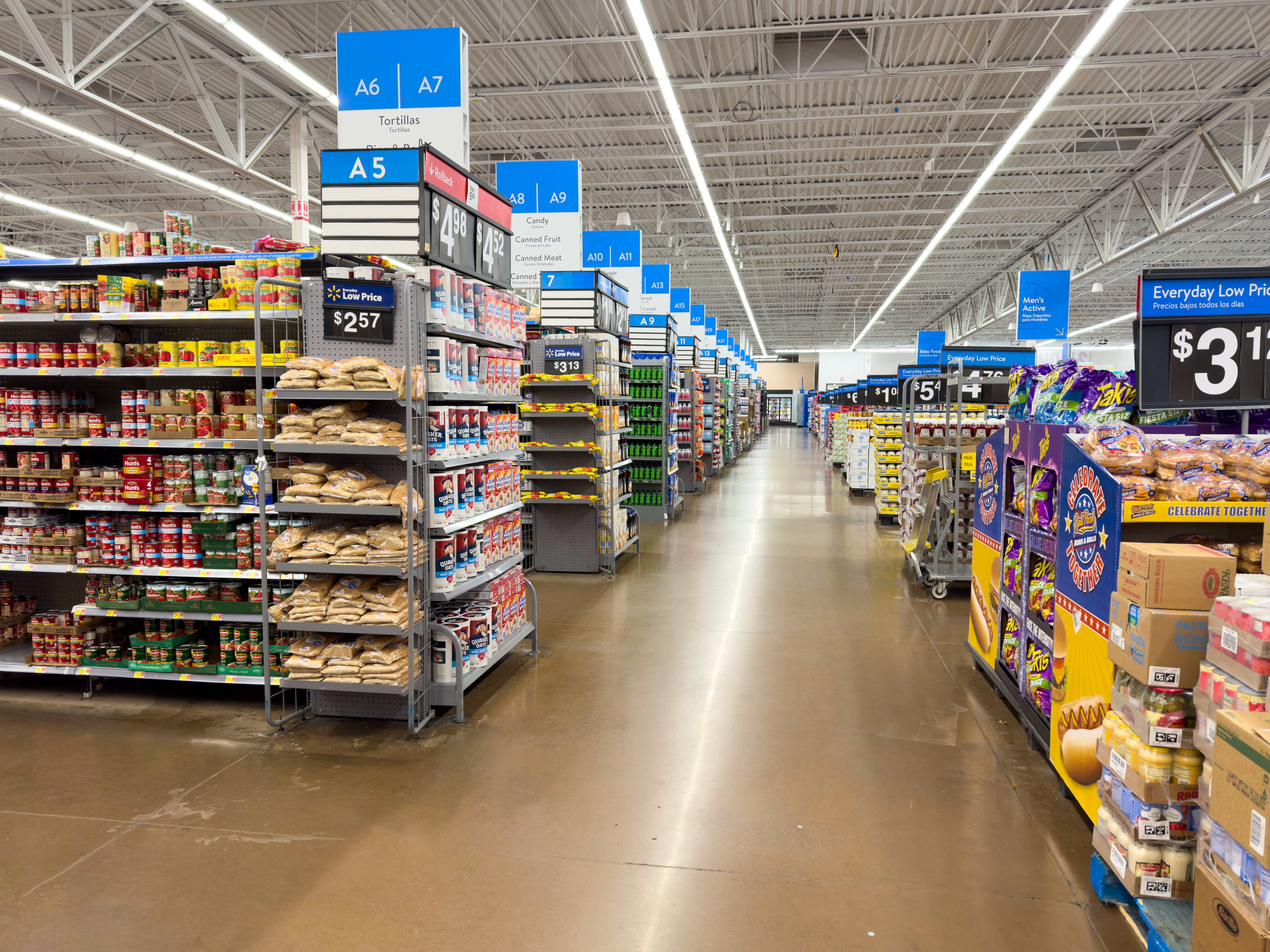 A wide aisle in a large retail store, featuring shelves stocked with various household items such as appliances, glue sticks, and cleaning products. The aisle is brightly lit with a "Home" section sign visible overhead.