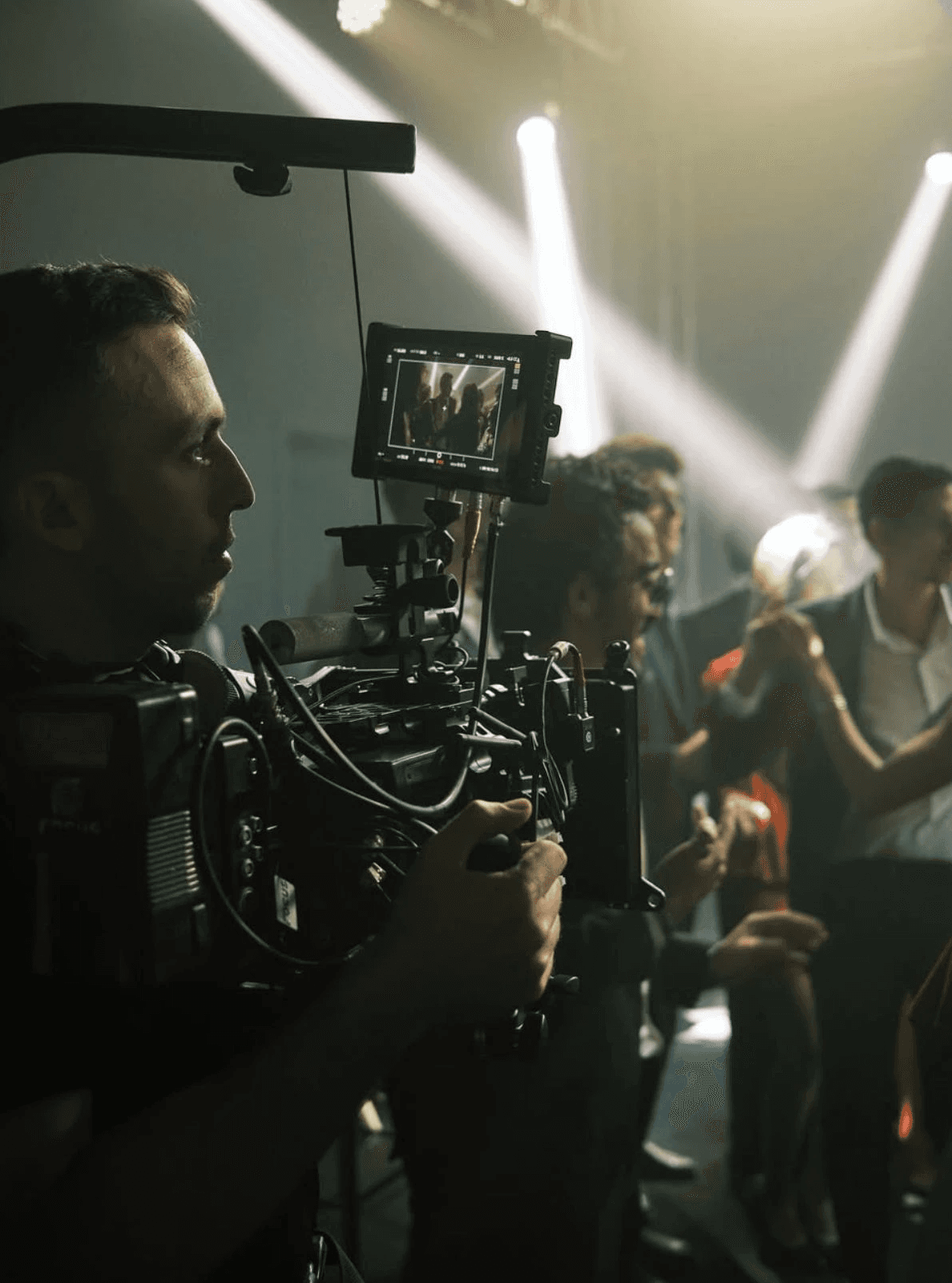 A cinematographer films a scene with a professional camera.  Bright stage lights illuminate the background where other people are visible.
