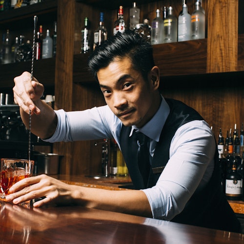 Bartender in a vest and tie stirs a drink with a metal rod at a wooden bar, with shelves of liquor bottles in the background.