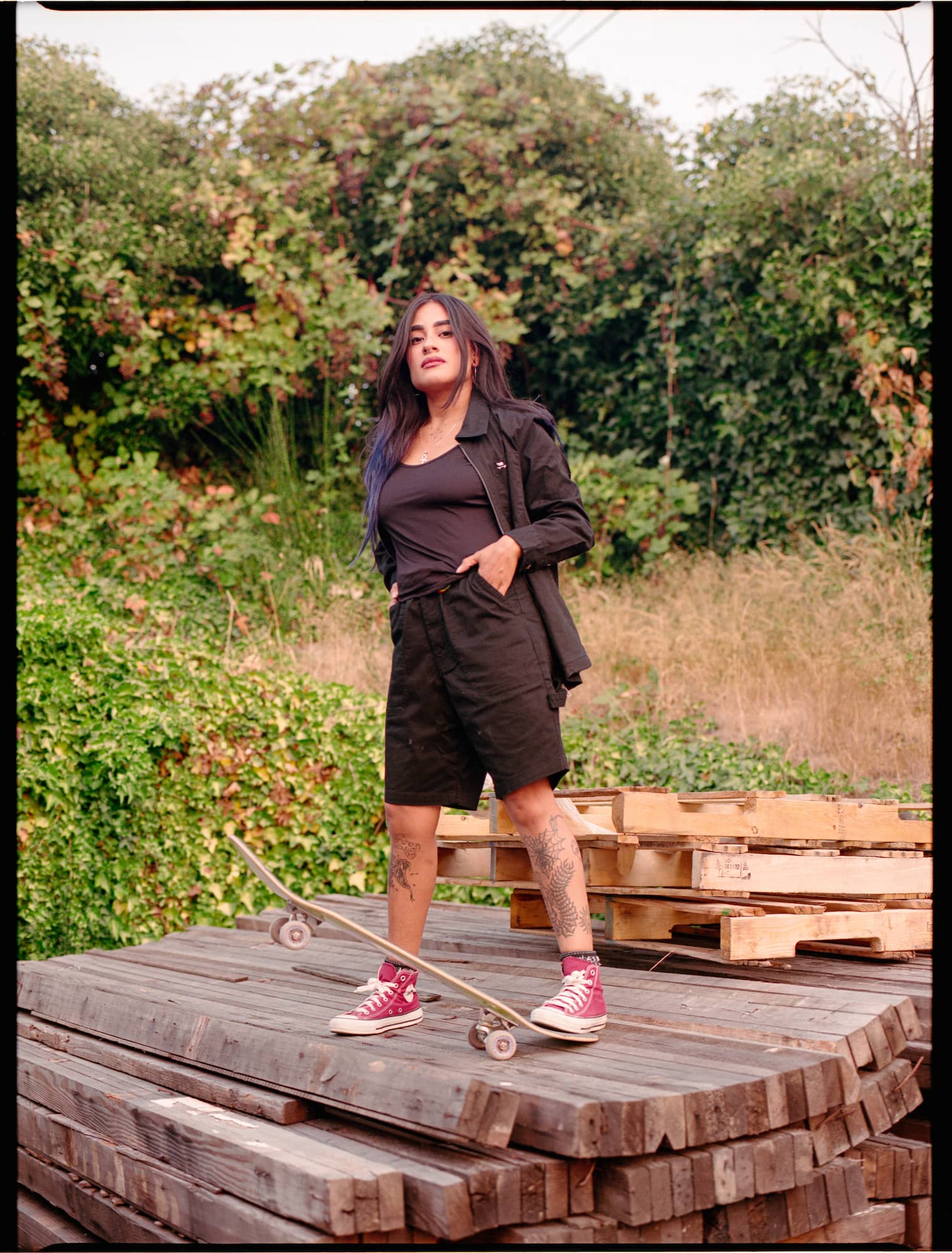 A person in a black outfit and red sneakers stands confidently on stacked wooden planks with a skateboard. Background features lush greenery.