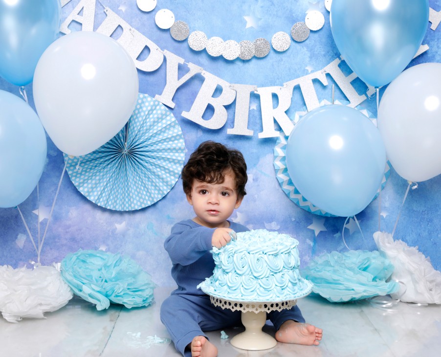 A baby boy is touching a blue floral cake in a blue background, with blue-and-white balloons. There is a “HAPPY BIRTHDAY” banner behind him.