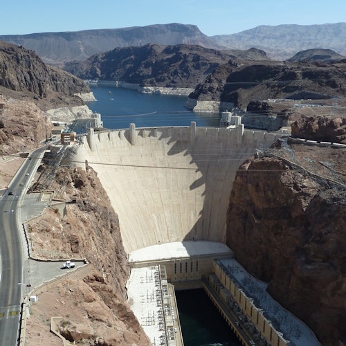 一座横跨峡谷的大坝，背后是水域，四周是崎岖的地形和道路。背景中可见山峦。