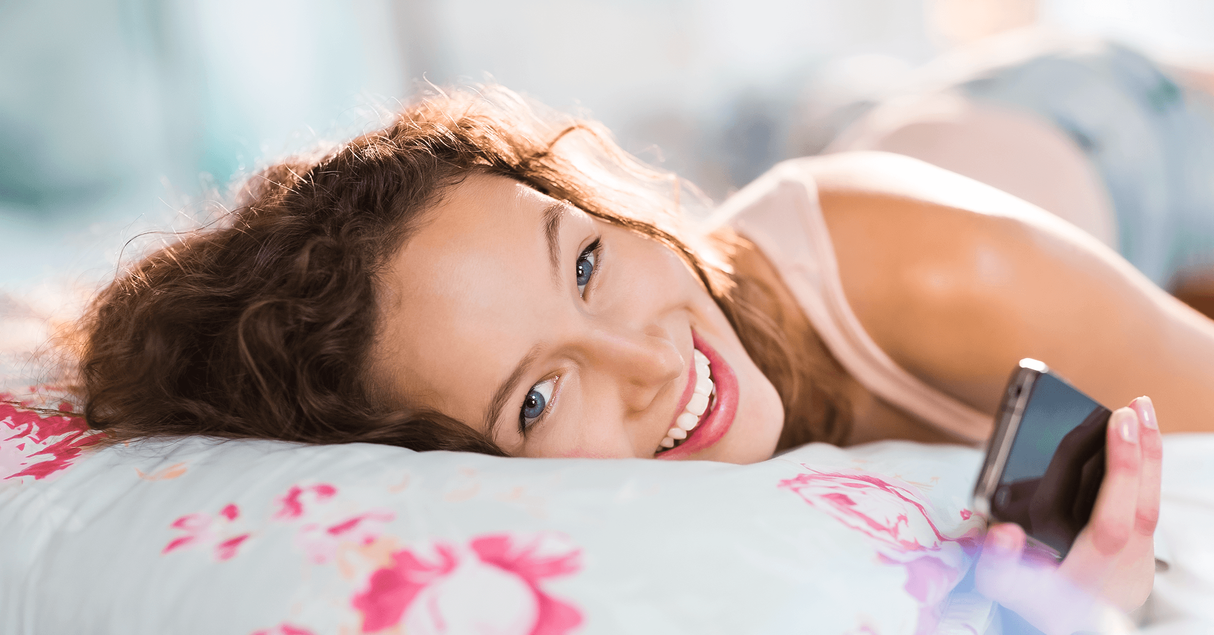 En kvinna ligger i sängen med sin mobil i handen medan hon tittar och ler mot kameran.