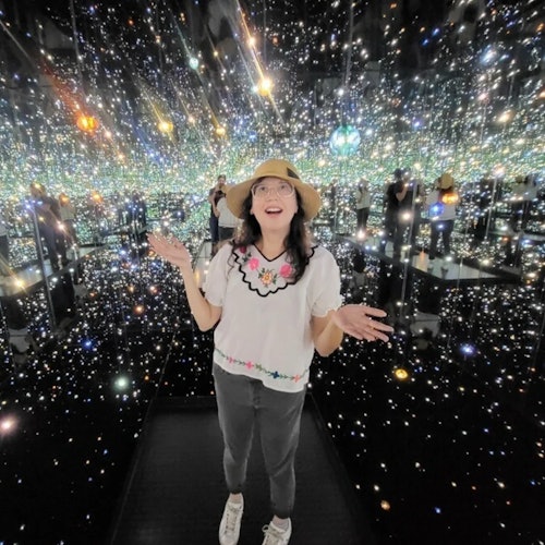 A woman stands in a room filled with mirrored walls and twinkling lights, wearing a hat, glasses, floral top, gray pants, and sneakers.