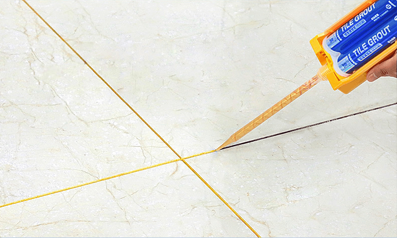 A worker is applying yellow tile grout to the joint between four marble tiles using a grout gun. The grout fills the gap neatly, providing a clean, smooth finish between the tiles.