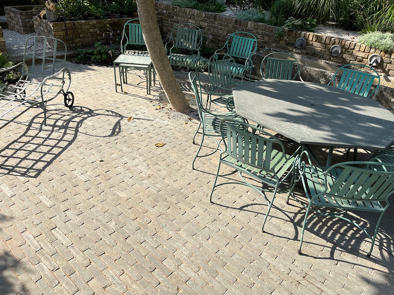 A sunny outdoor patio with stone flooring, metal tables, and chairs surrounded by greenery.
