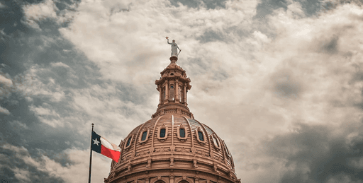 The Texas State Capitol building under a partly cloudy sky with the Texas flag in the foreground.
