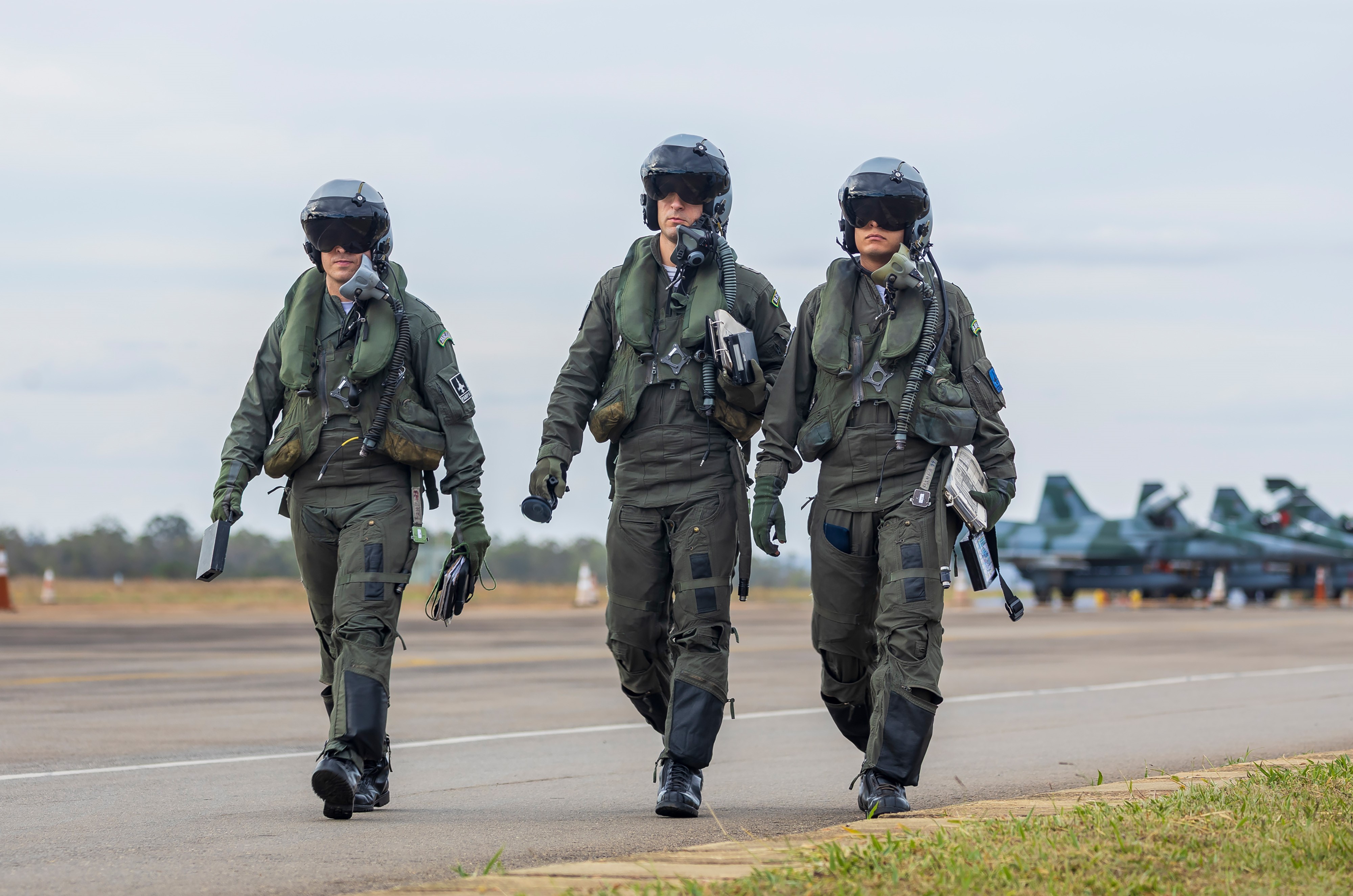 Três pilotos da Força Aeria Brasileira caminhando juntos com três aviões atrás.