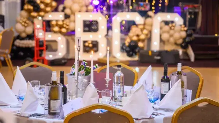 Elegant banquet hall with a round table set for dining, featuring white napkins, wine bottles, and candles. Large lit "SBCC" with balloons in the background.