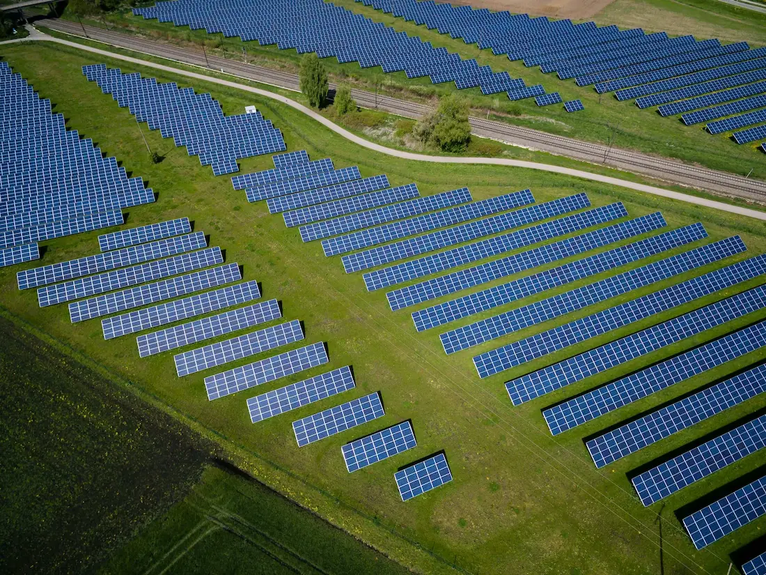 Aerial view of a renewable energy solar farm with rows of panels in a green field next to railway tracks.