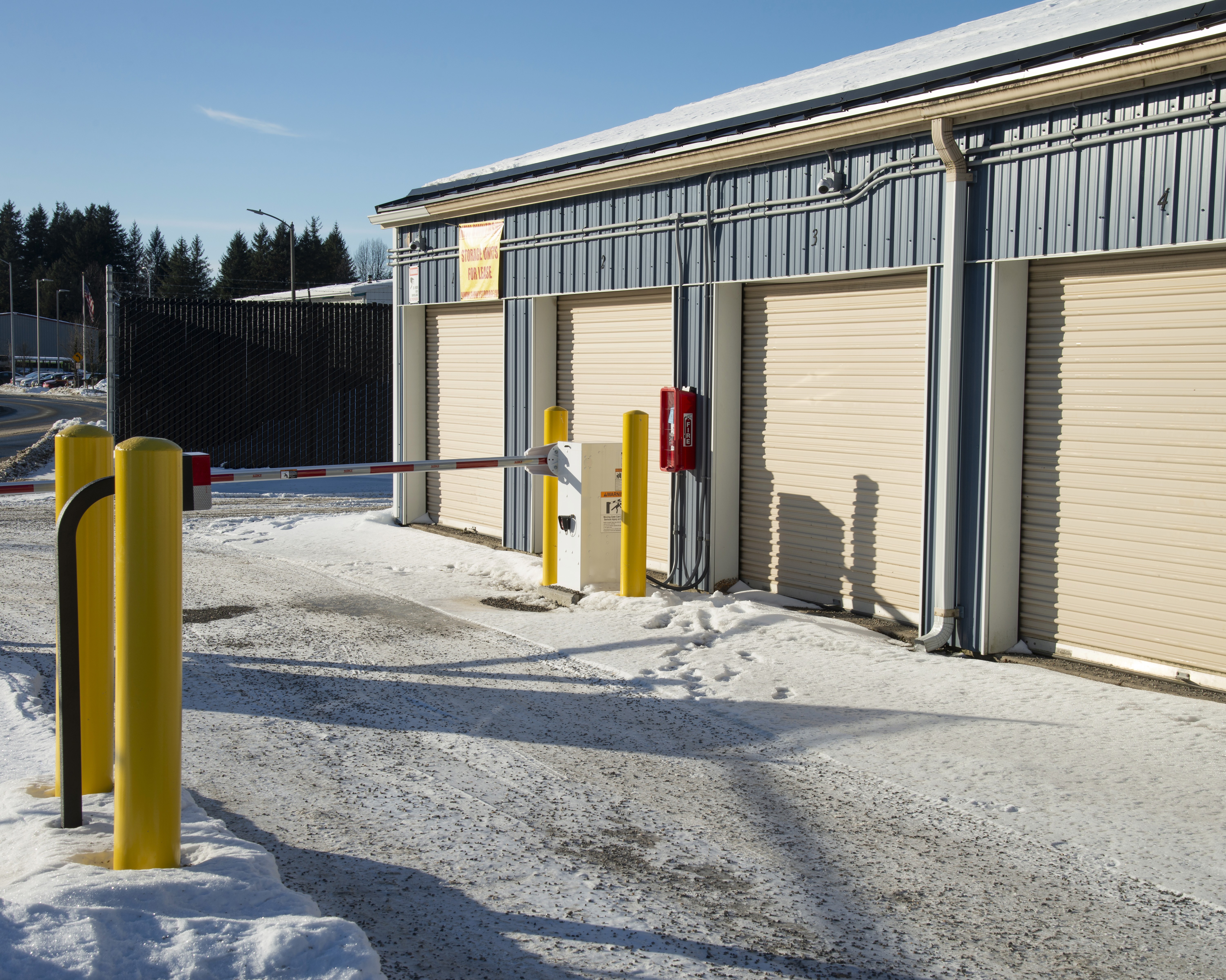 Exterior access point of a self-storage facility in winter, showing beige roll-up doors, a yellow bollard-protected keypad entry system, a security camera, and a gate arm, with snow-covered ground and a perimeter fence visible in the background.