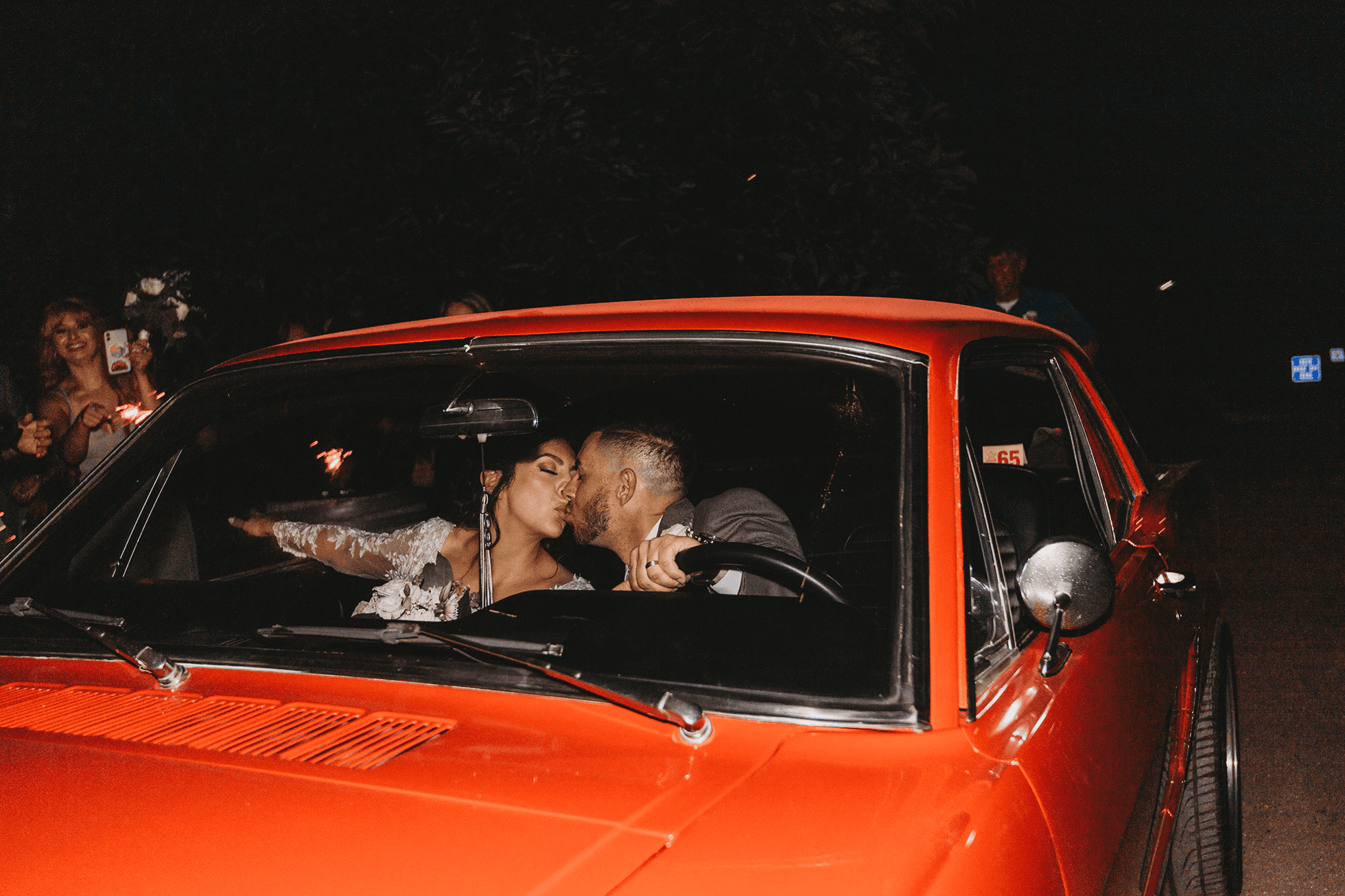 Bride and groom kissing inside red Mustang after sparkler exit