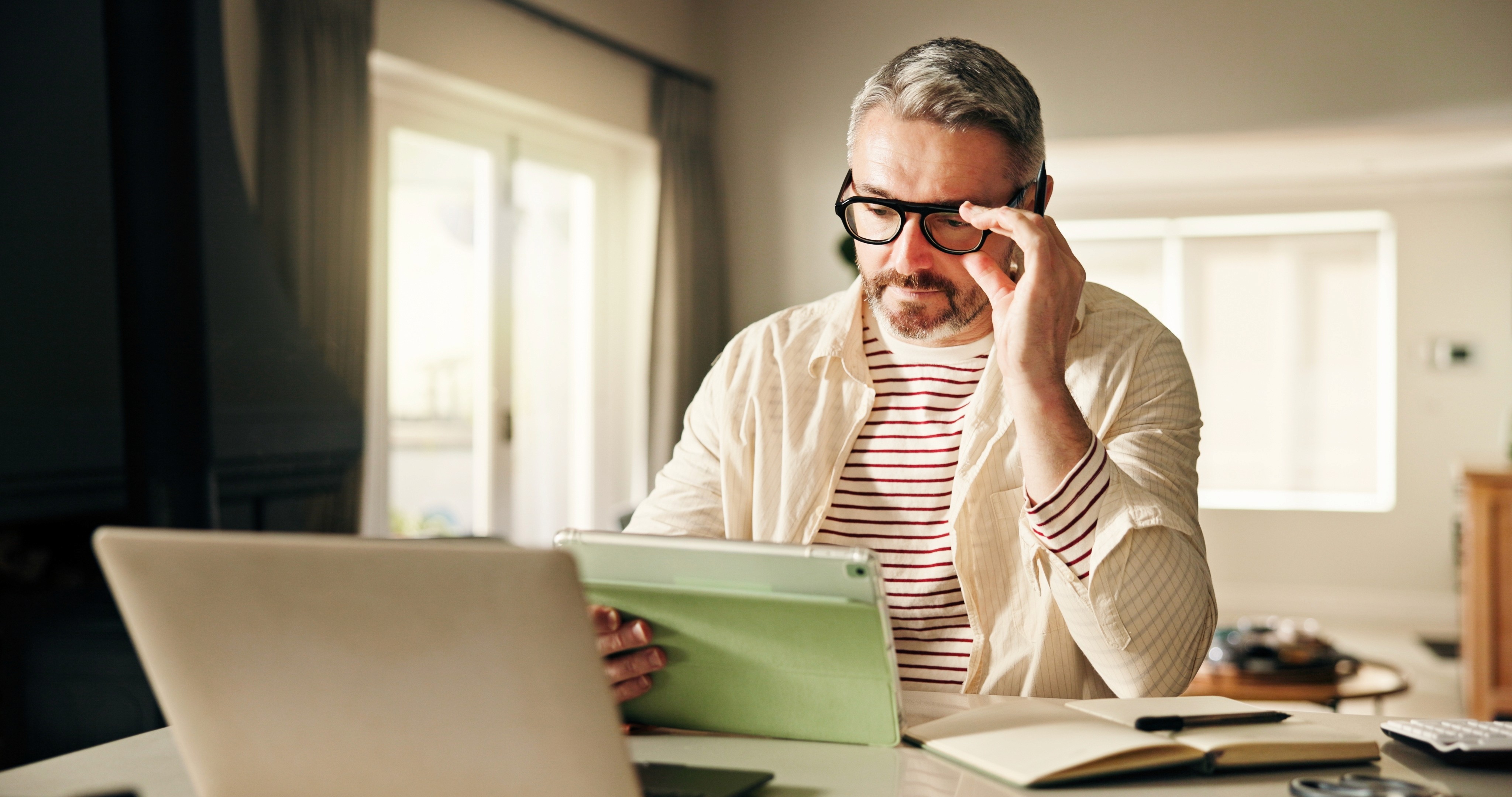 Man in glasses wearing a green shirt, writing in a notebook at a speckled table with a cup of coffee nearby, engaged in conversation.