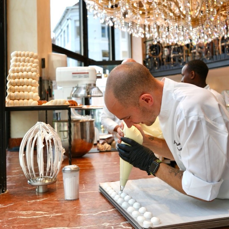 Un pâtissier dresse à la poche des dizaines de meringues identiques sur une plaque, sous les lustres de la boutique