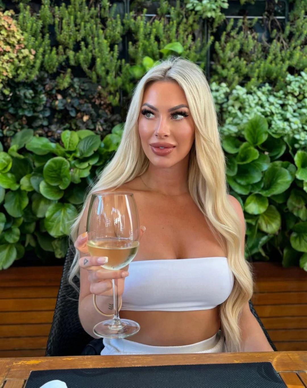 Topless waitress Vanessa sitting outdoors with a glass of wine, wearing a white top for an Hourglass Gold Coast promotional photo.