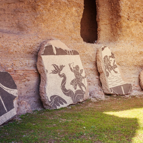 古老的石头马赛克上描绘着神话人物，靠在长满草的区域上的一面老化砖墙上。