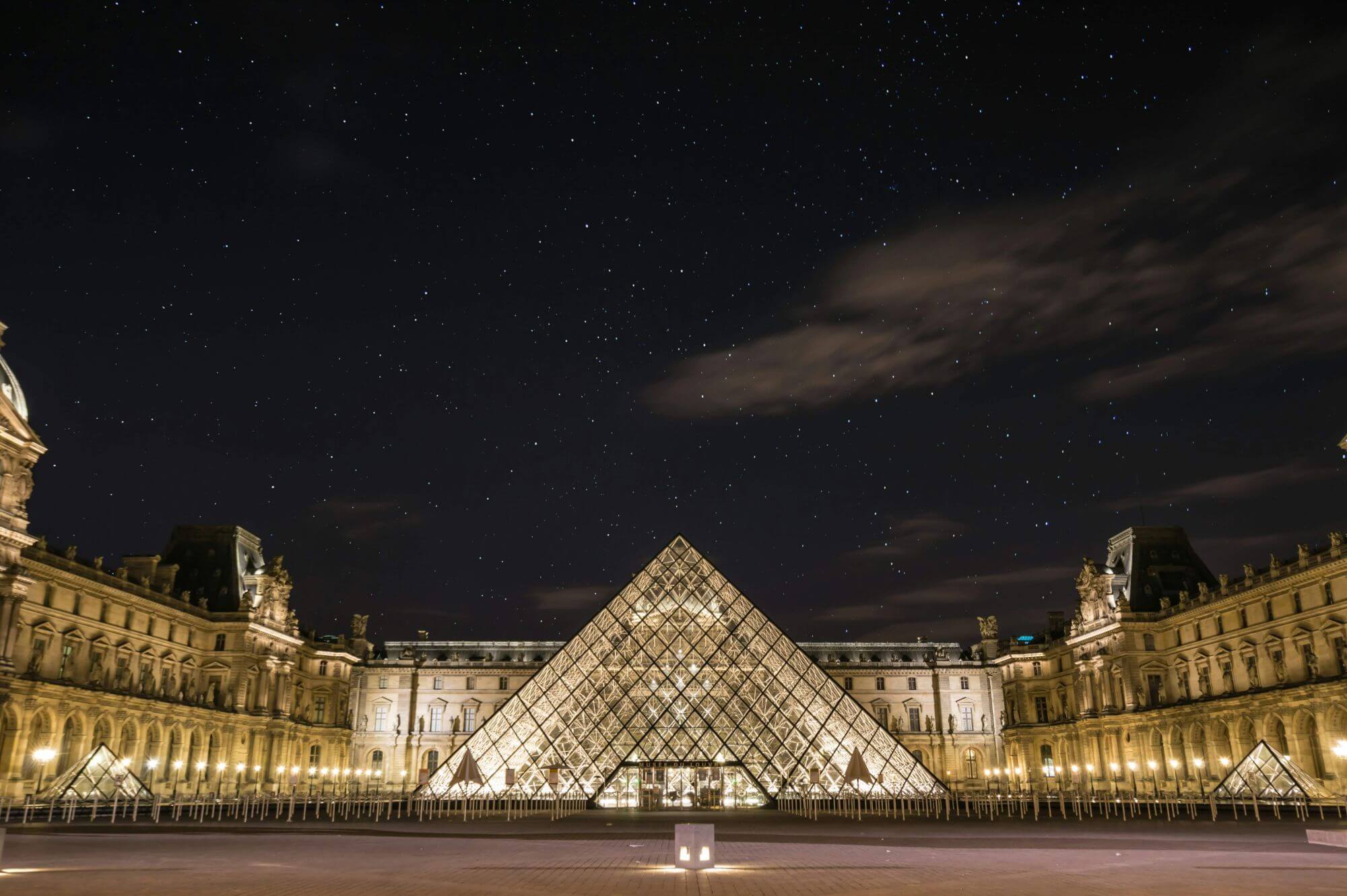 The image shows an illuminated glass Pyramid at night, framed by the historic palace buildings under a starry sky, conveying a serene, majestic atmosphere.