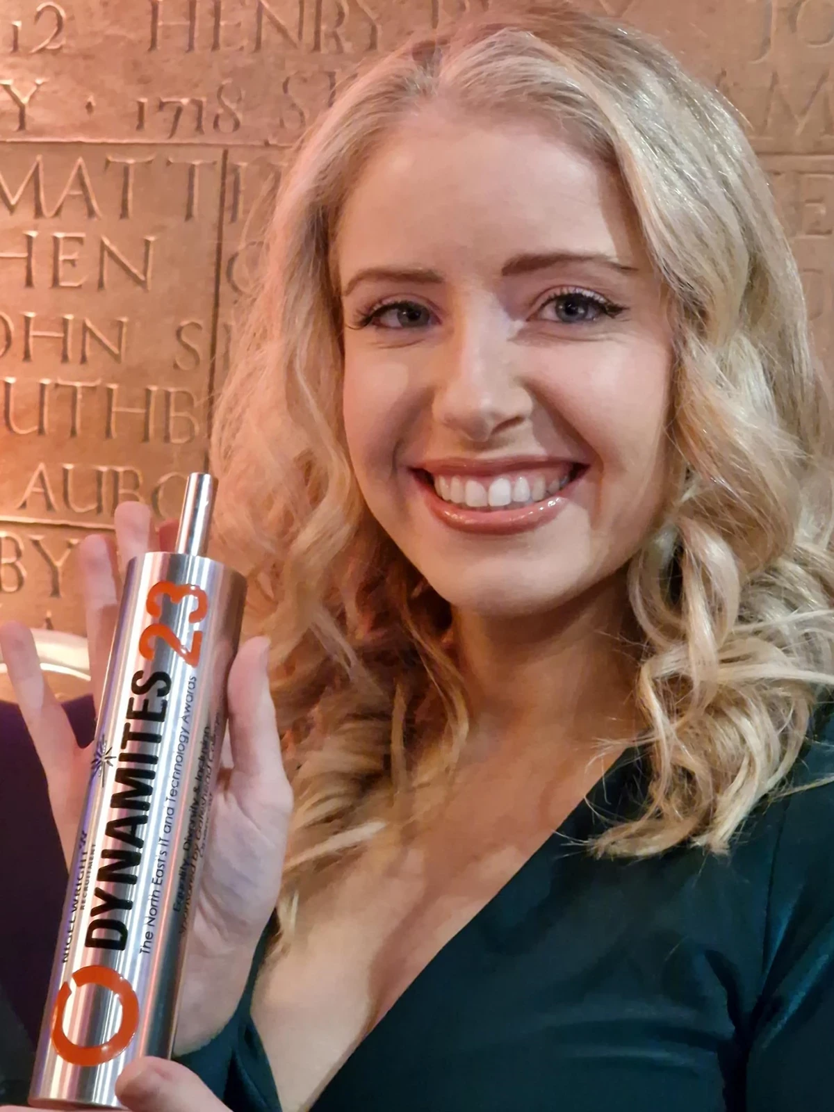 Person holding a silver Dynamites 23 award trophy with engraved text, standing in front of a wall with carved names.