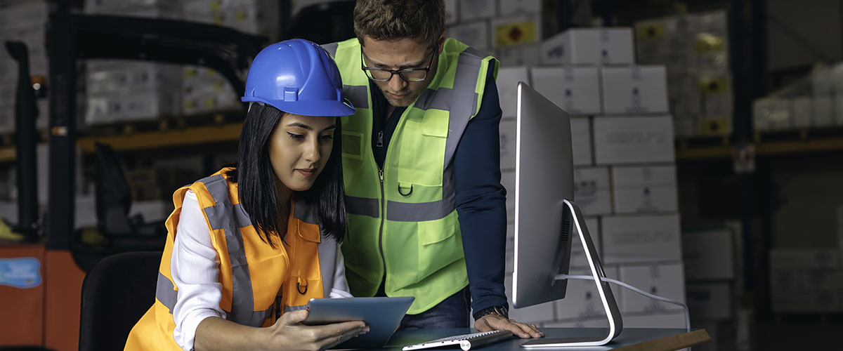 Two workers in a warehouse, one wearing a hard hat and safety vest, collaborating on a project.