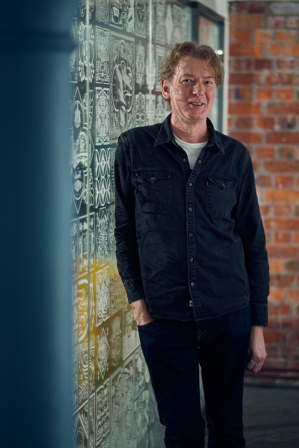 Tom Lawson standing on a stage under dramatic blue and purple lighting, wearing a light blue button‑up shirt. They are positioned near a lectern and microphone stand, holding their hands together as if mid‑talk. The dark background and spotlighting suggest a live presentation or speech at a professional event or conference.