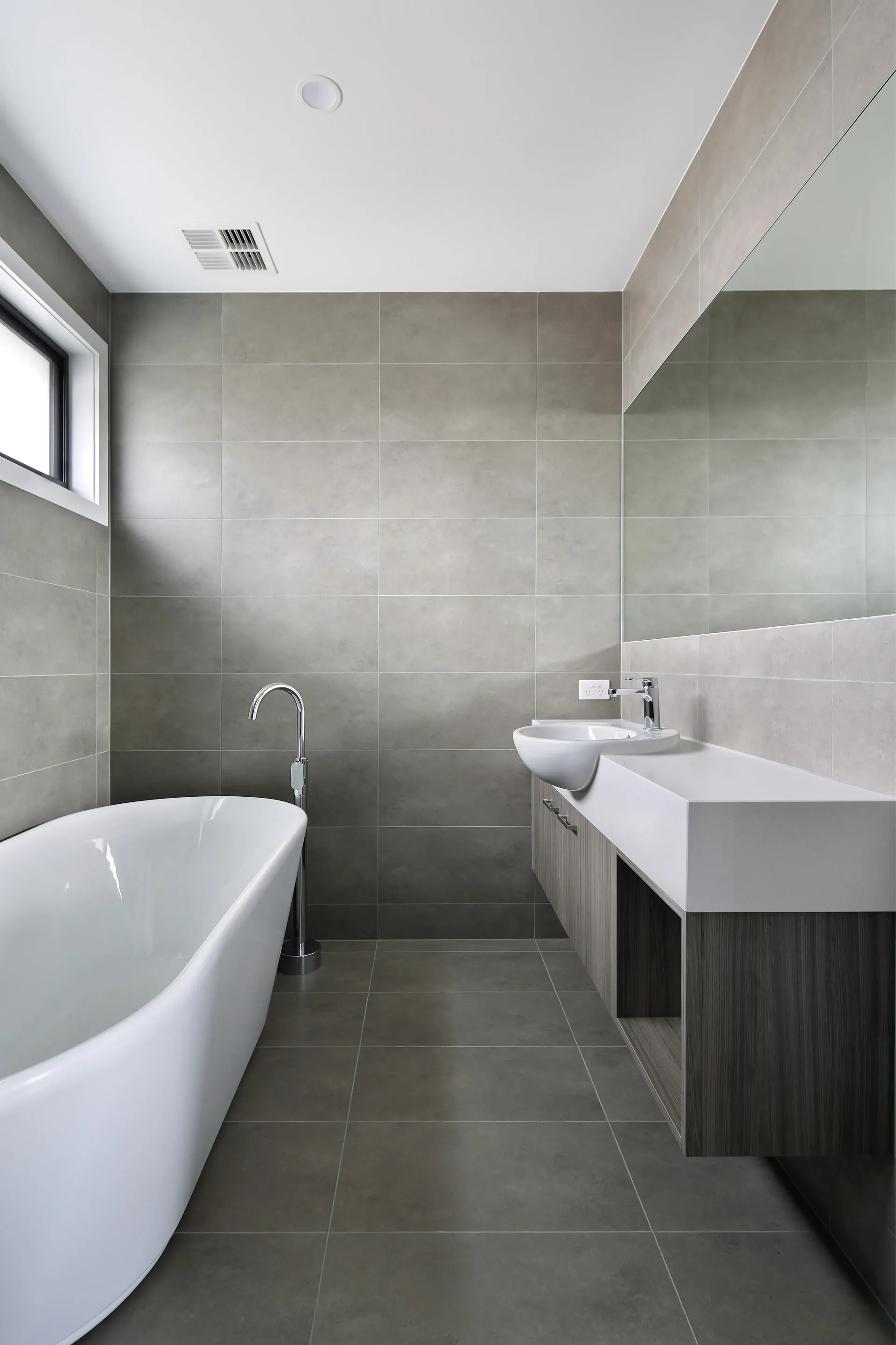Bathroom in large grey tiles with a freestanding white bath, floor-mounted chrome bath filler, a timber-veneer floating vanity with white basin and a frosted black-framed window