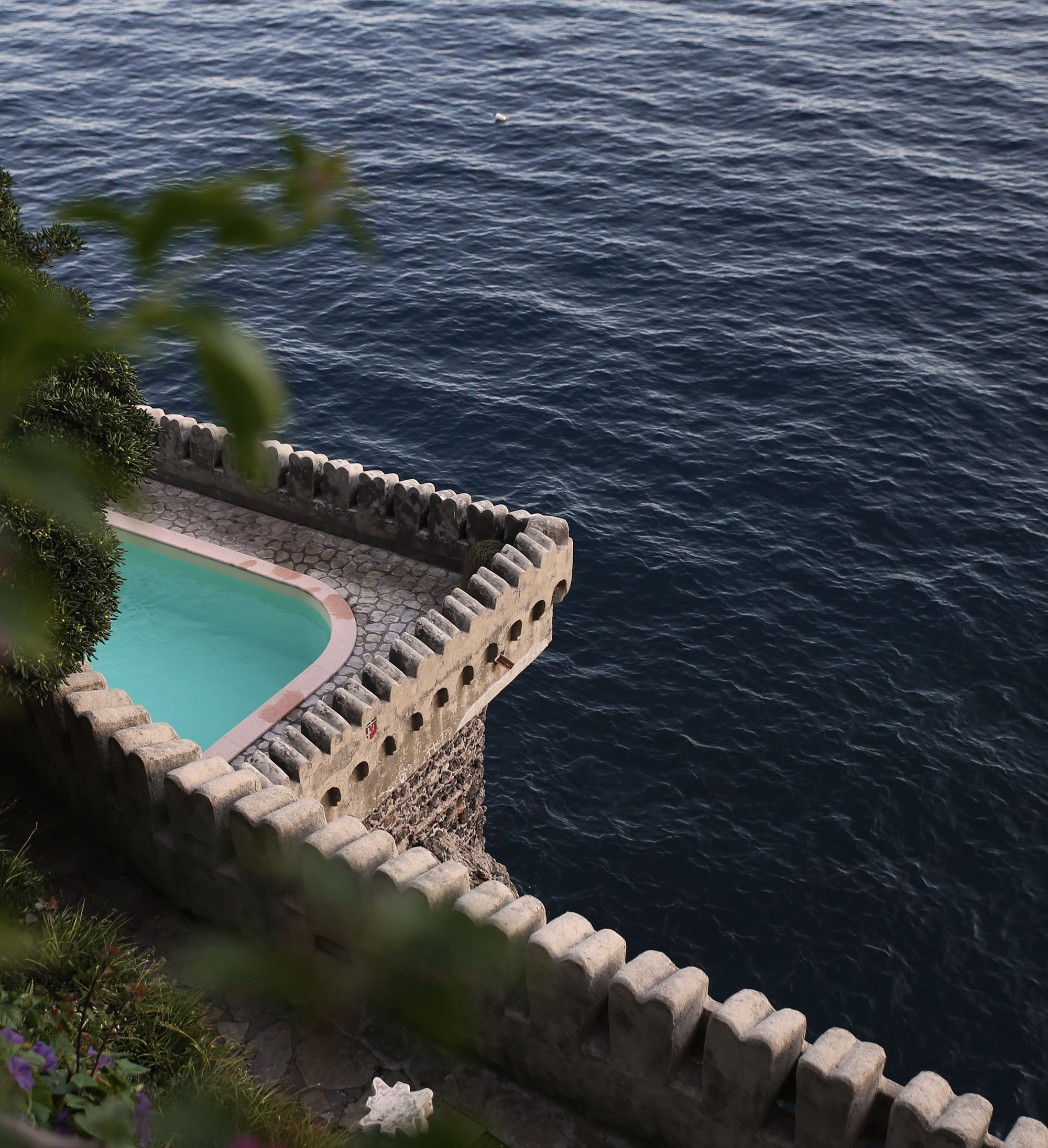 Aerial view of a cliffside infinity pool with castellated stone walls above the deep blue sea