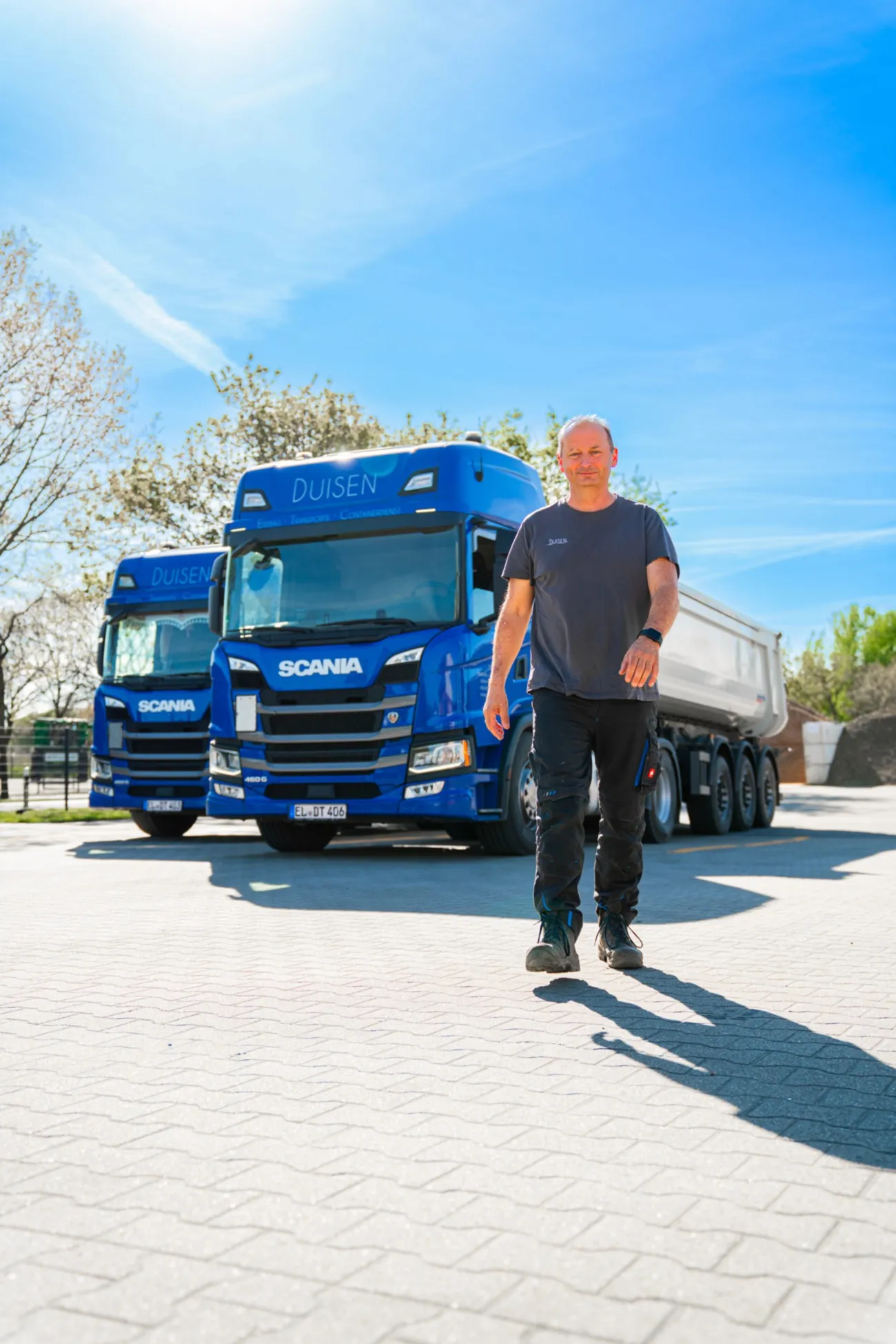Fahrer läuft vor zwei blauen Scania-Lkw „Duisen“ über den Hof