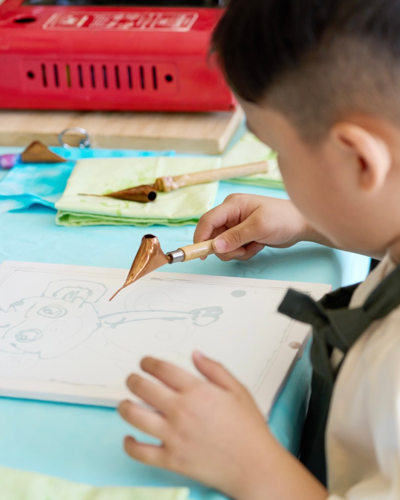 A child holding a traditional tool for batik painting, focused on their artwork on a piece of paper.