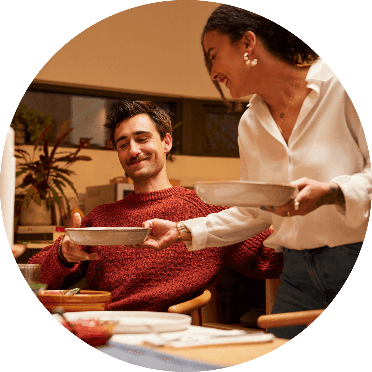 Woman serves a dish to a smiling man seated at a dining table in a cozy home setting