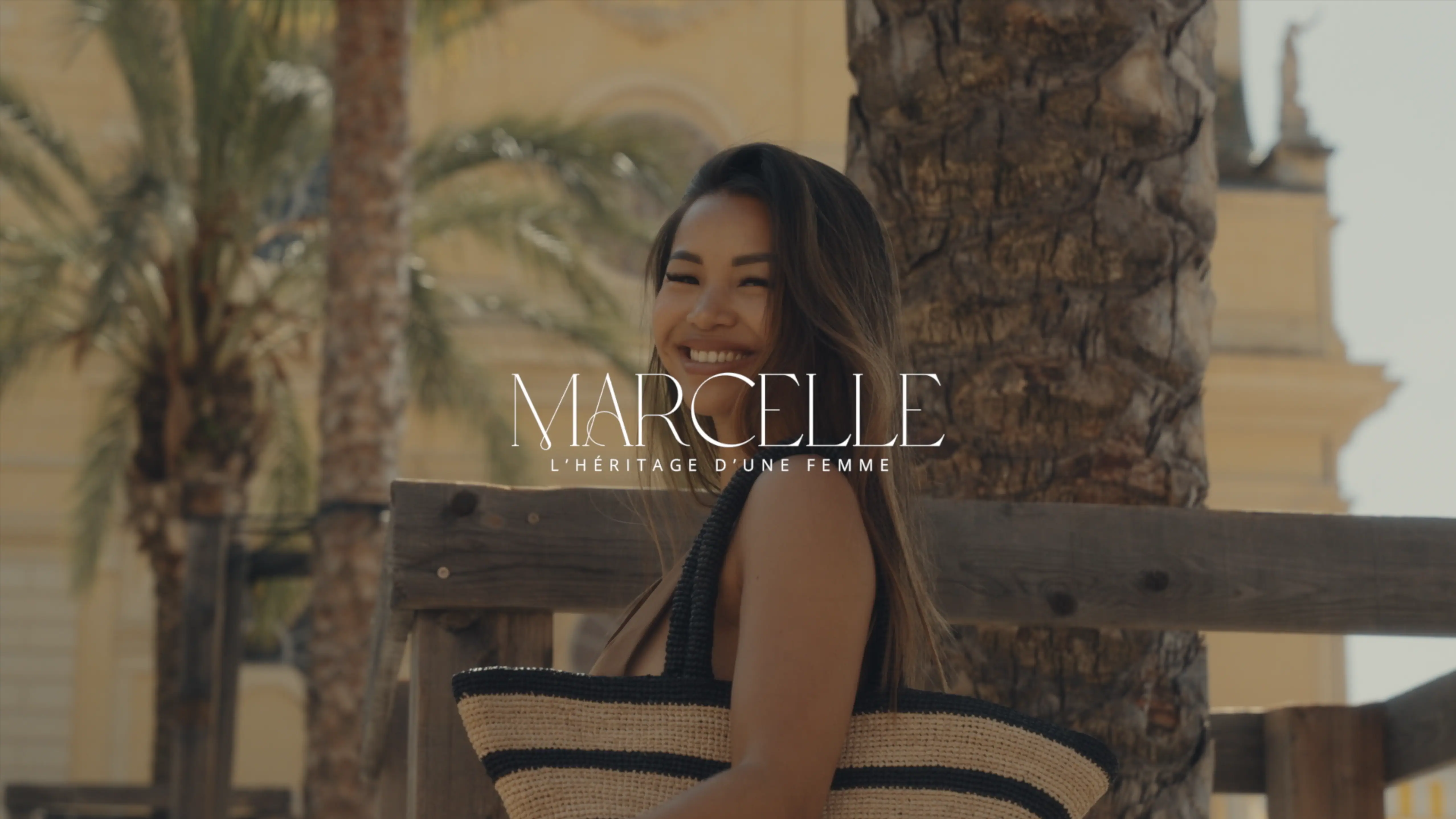 A woman smiles while holding a large straw bag, standing outdoors in a sunny location with palm trees and elegant architecture in the background.
