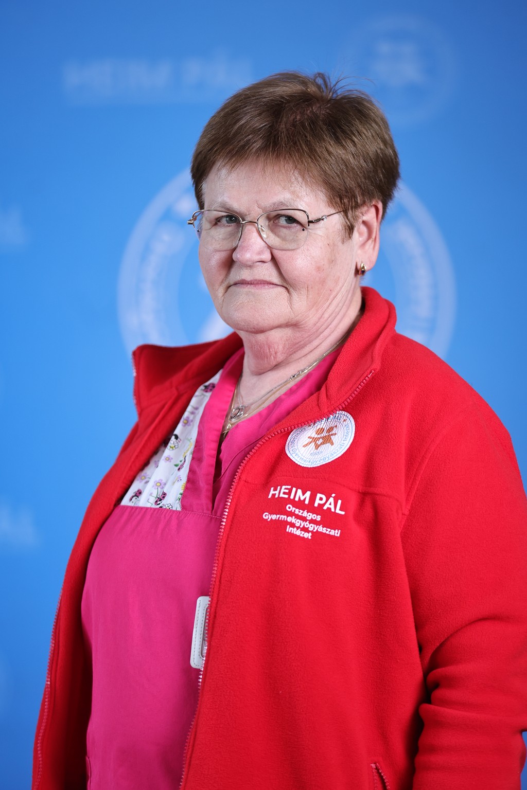 Sötét hajú nő mosolyogva áll egy hivatalos, kék háttér előtt, piros ruhában.