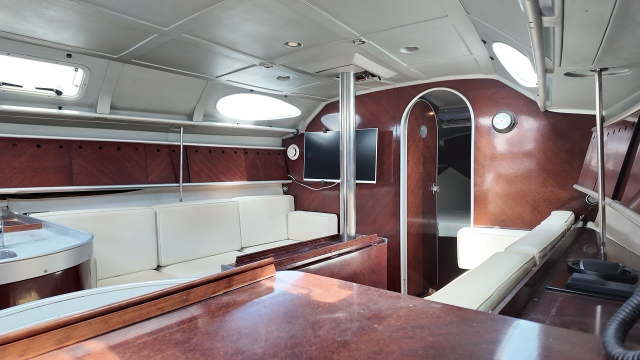 Interior view of the Beneteau First 38.5's saloon, showing curved cream cushioned settees, dark mahogany joinery, a wall-mounted TV, and an arched doorway leading to the forward cabin.