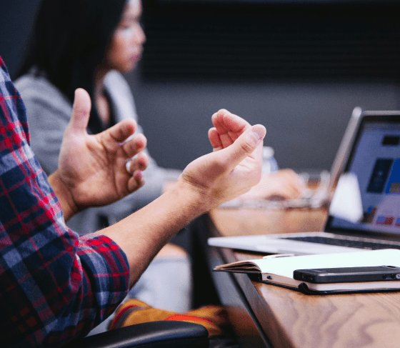 A designer’s hand gesturing while discussing design concepts with a stakeholder, with an open laptop in view.