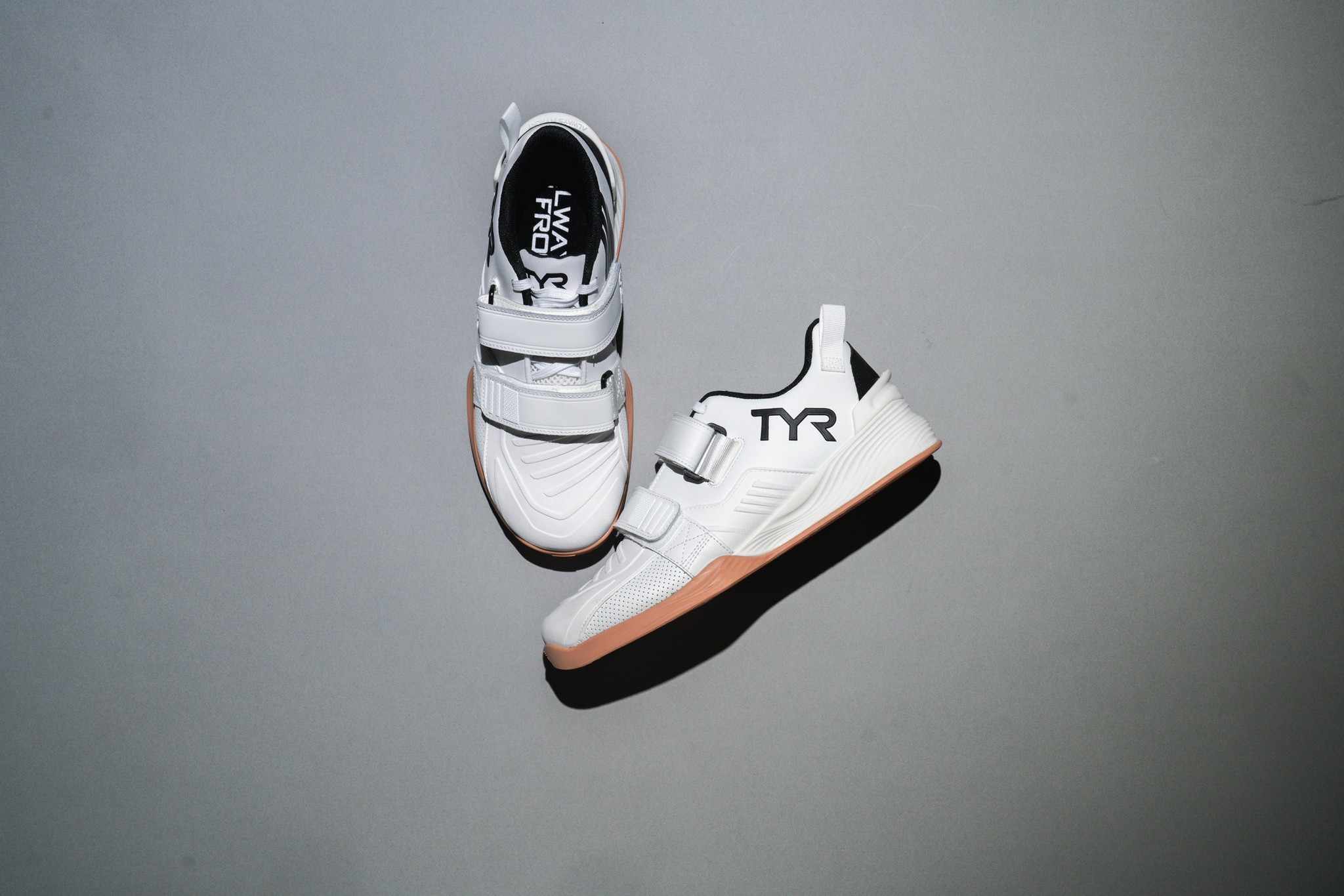 A pair of white TYR training shoes with gum soles displayed on a gray background, styled for product photography.