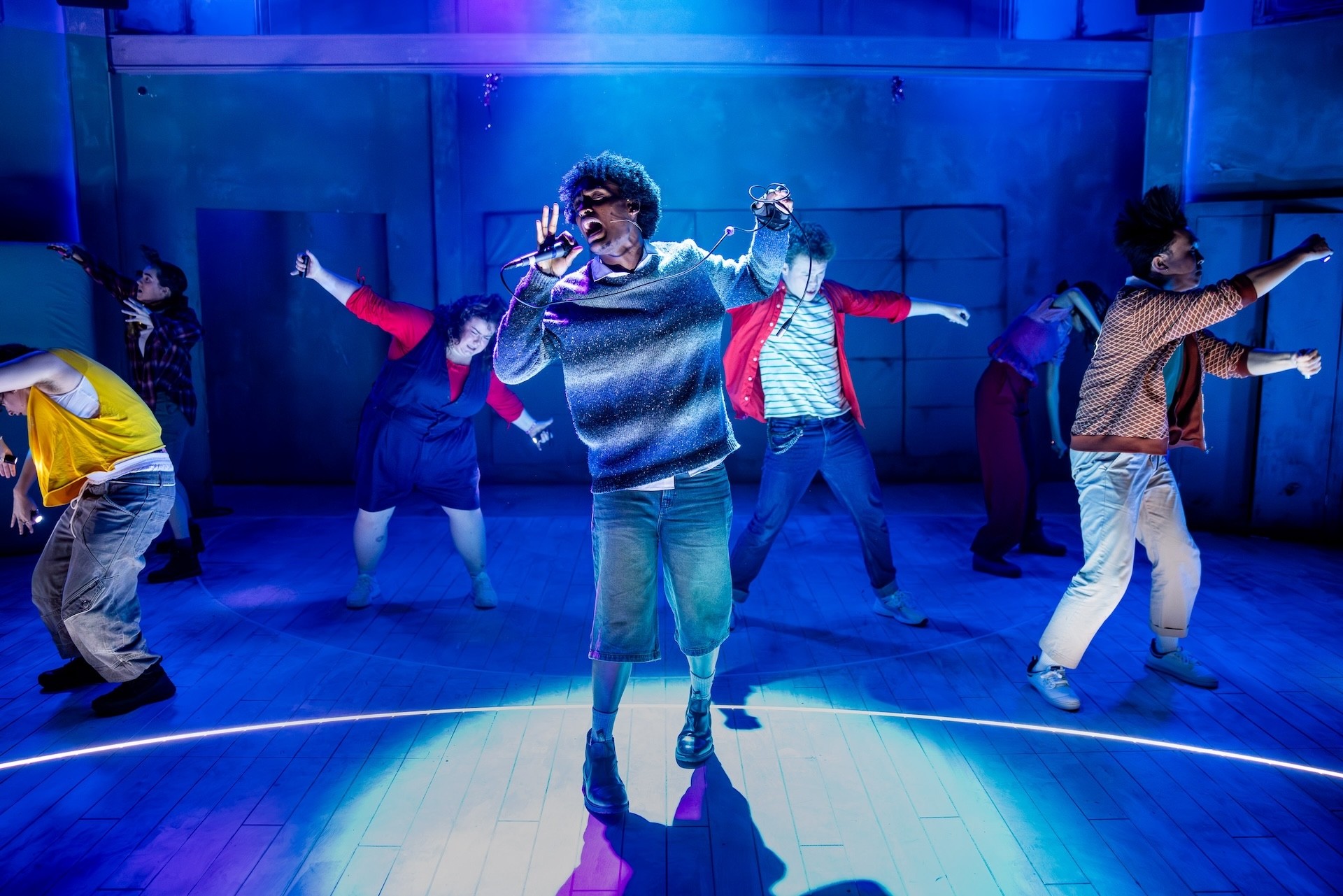 A group of young performers dance energetically on a stage washed in blue light. A performer at the centre sings passionately into a microphone, while the ensemble moves in dynamic poses around them. The scene is vibrant and expressive, with colourful costumes and dramatic theatrical lighting.