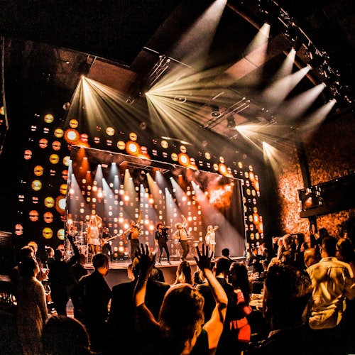 Sebuah band tampil di atas panggung yang terang benderang dengan lampu-lampu warna-warni, sementara penonton mengangkat tangan mereka dan bersorak.