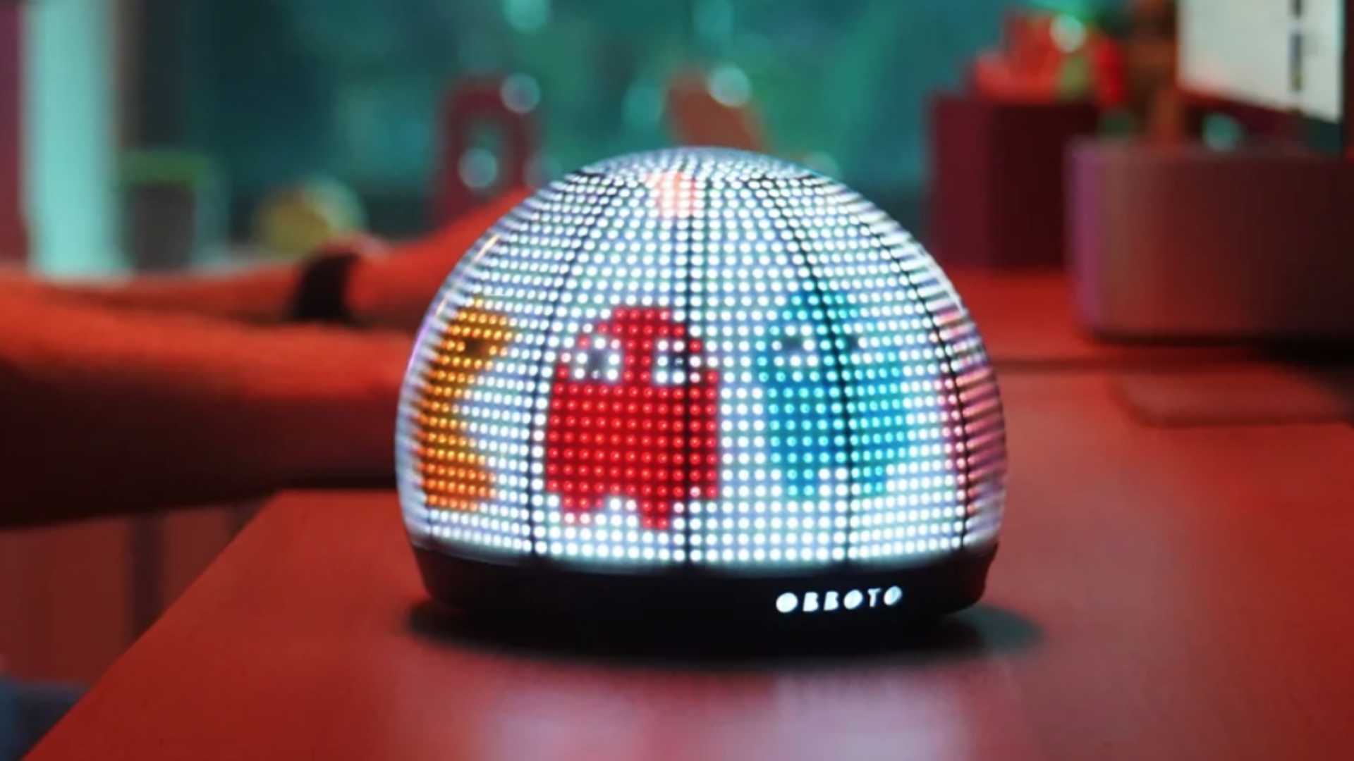 Close-up of the OBBOTO Glowbot sitting on a desk, a spherical smart lamp with a pixelated LED display showing a red character, representing the shift to ambient AI companion devices.