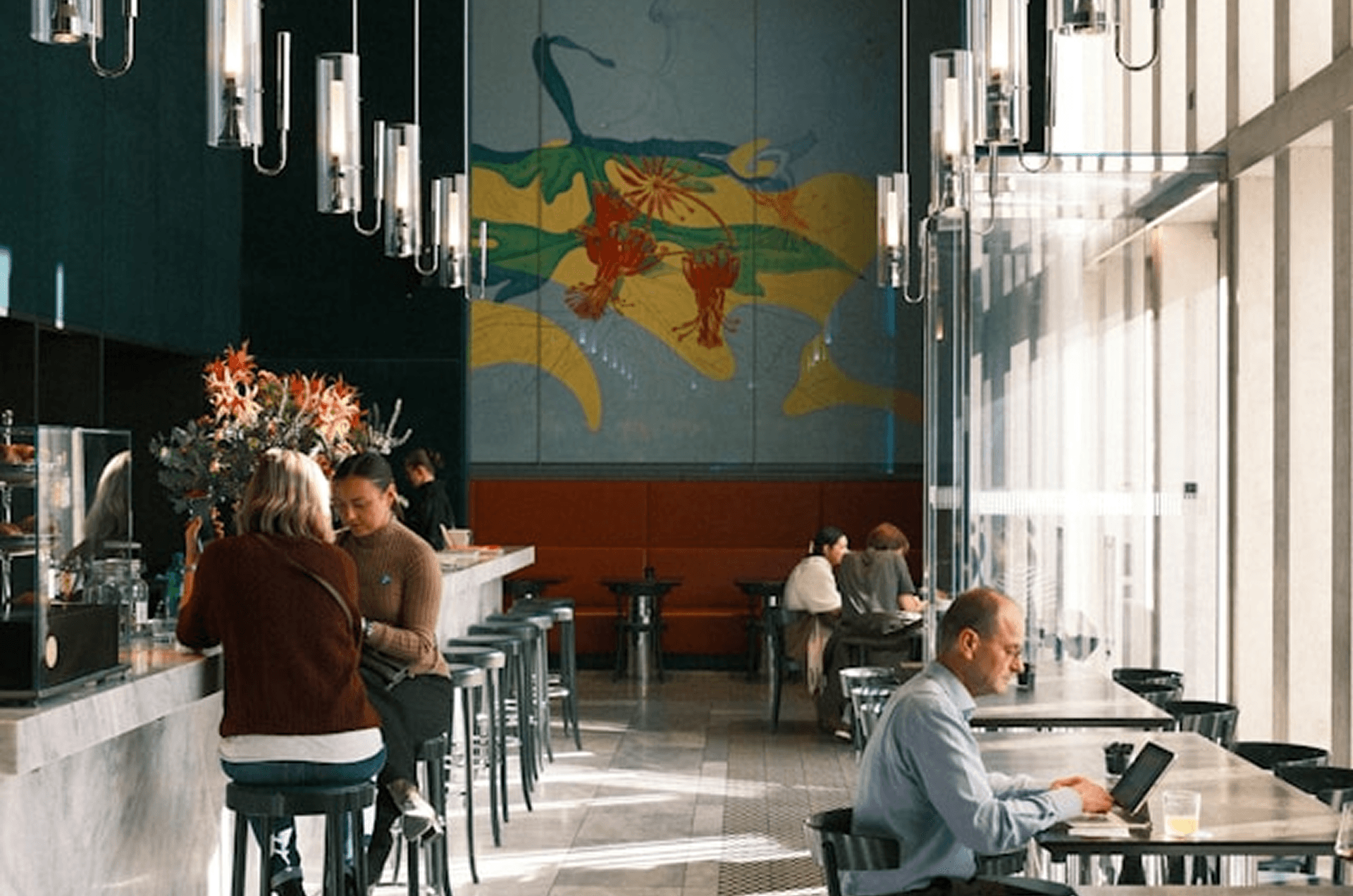 Modern café interior with natural light, featuring people working and socializing at tables and a bar, pendant lighting overhead, and a colorful wall mural—ideal for coworking, remote work, lifestyle, and urban coffee shop settings.