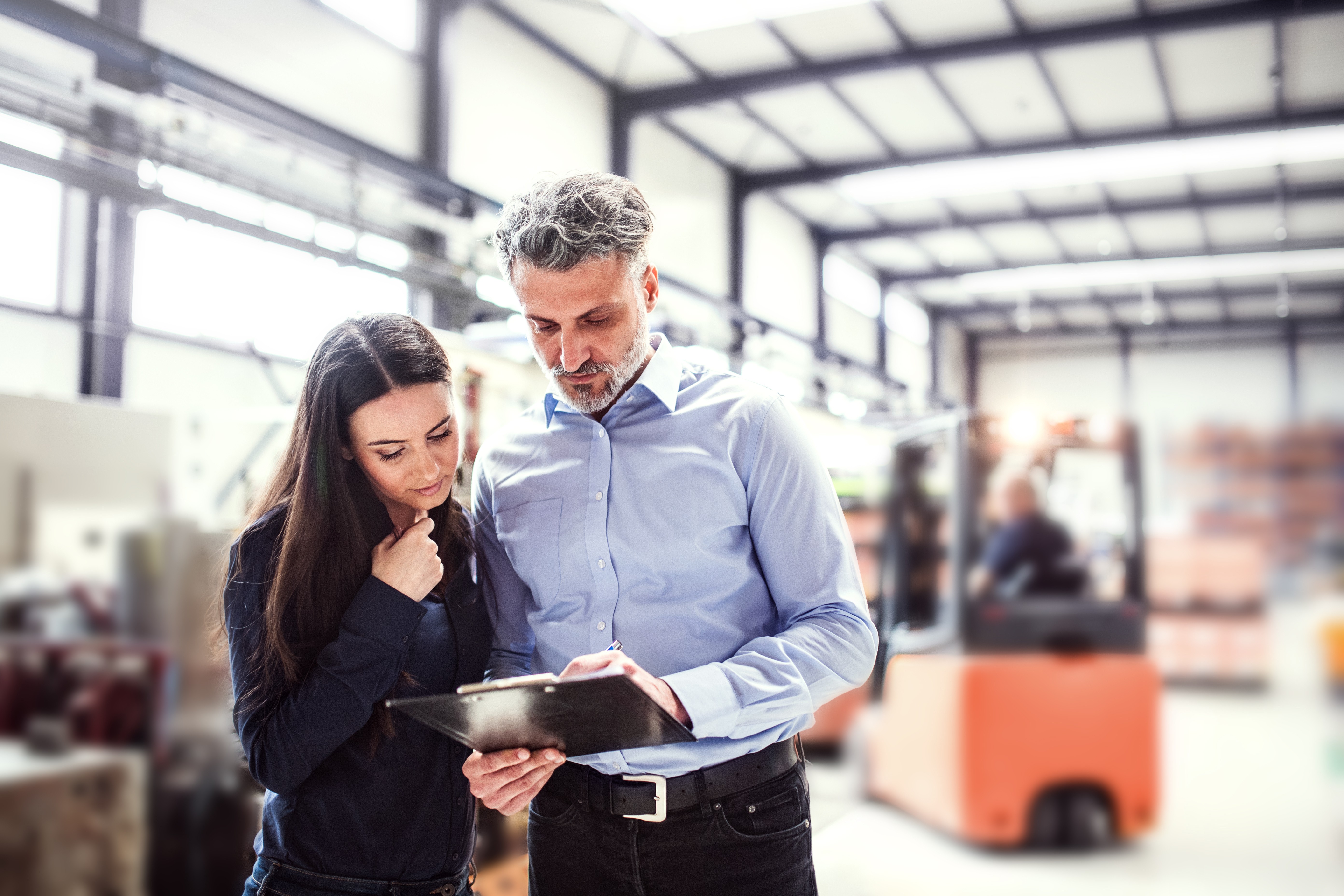 Zwei Mitarbeitende in einer Lagerhalle schauen gemeinsam auf ein Klemmbrett und besprechen Notizen.