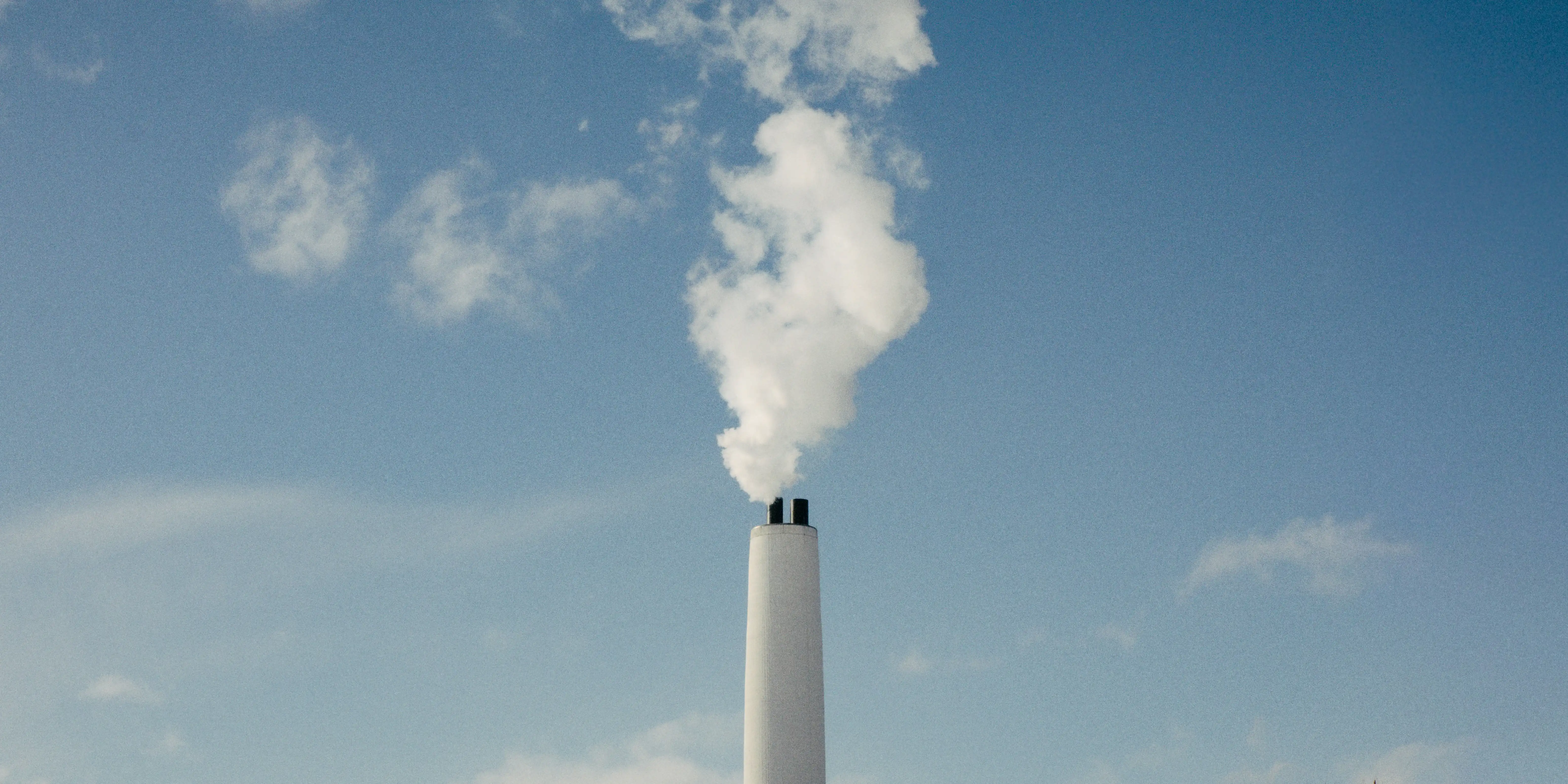 A tall white industrial chimney emitting a thick plume of smoke into a clear blue sky.