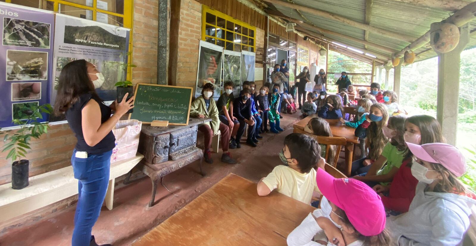 Educadora fala para alunos de máscara em varanda com painéis de fotos, durante visita escolar