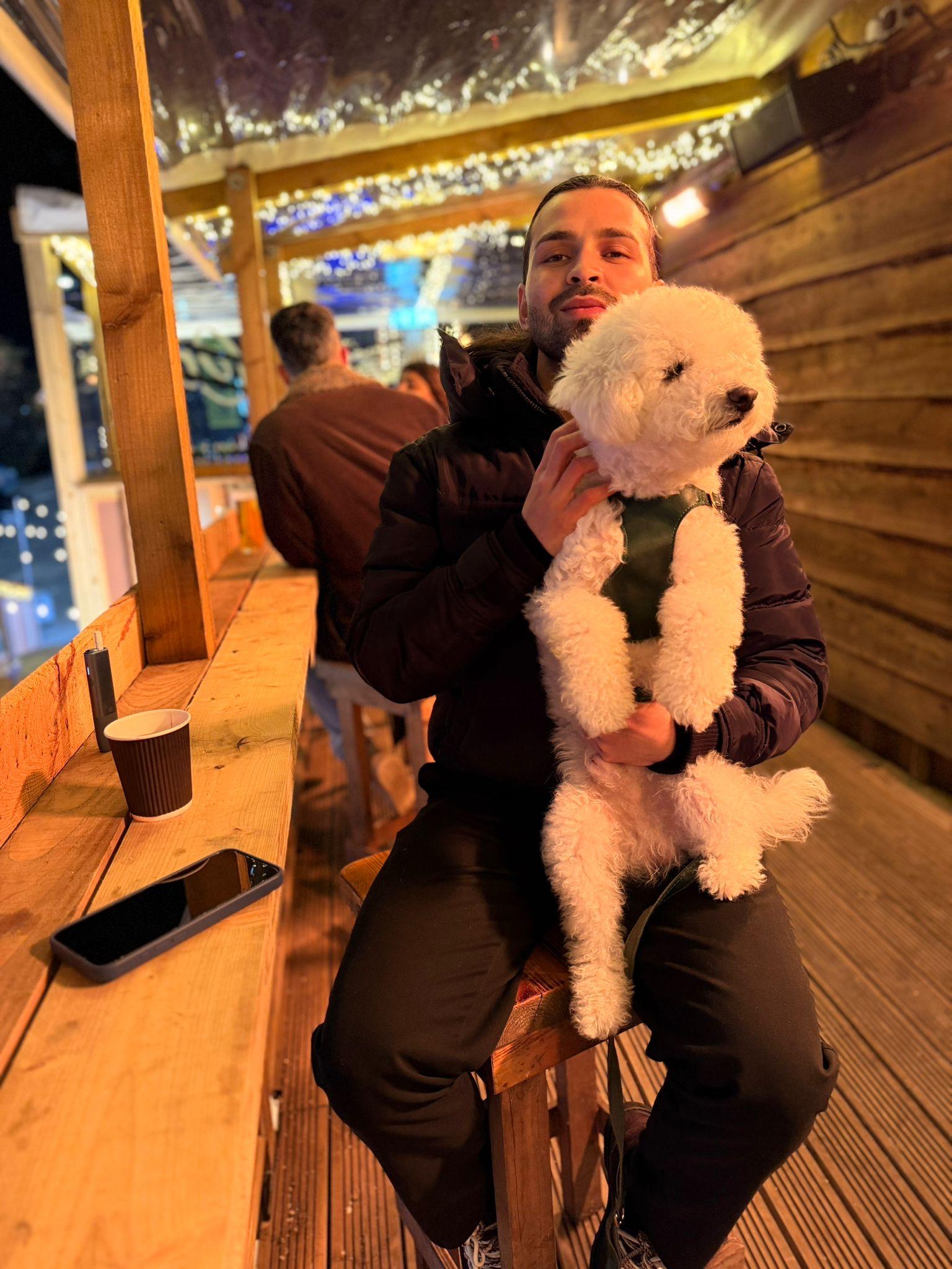 A man sitting at a wooden outdoor café in the evening, warmly dressed, holding a fluffy white dog on his lap, with a to-go coffee cup and a smartphone on the table beside him.