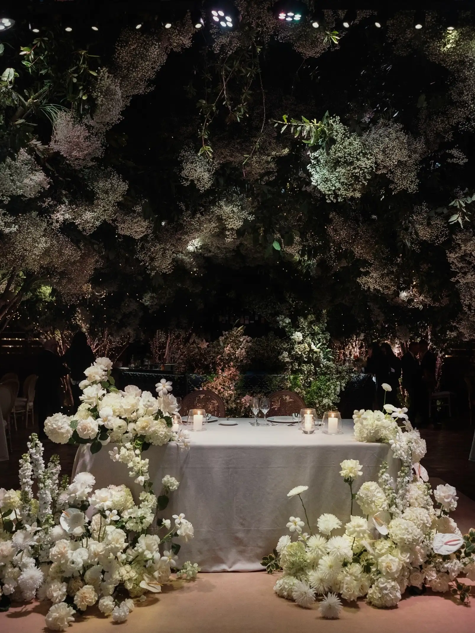 Mesa nupcial rodeada de flores blancas y follaje, bajo una atmósfera cálida y romántica.