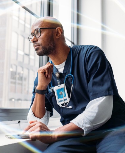 A male doctor in blue scrubs looking out a window while using a smartphone, symbolizing healthcare staff scheduling and clinical time management for hospitals.