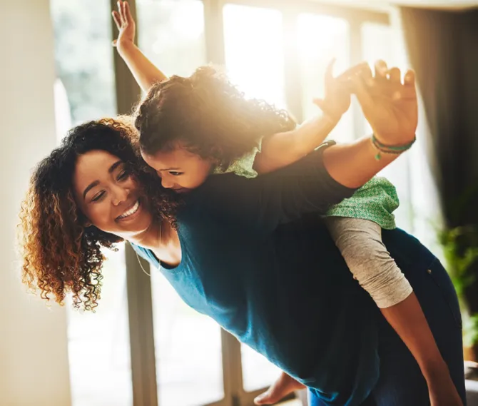 Happy mother giving her young daughter a piggyback ride indoors with sunlight streaming through the windows.