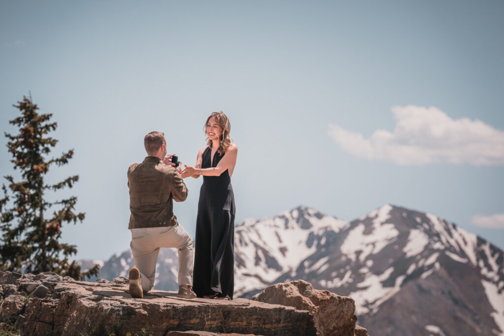 Romantic mountain proposal with partner kneeling and holding a ring box on a cliff, captured by a Colorado wedding photographer.