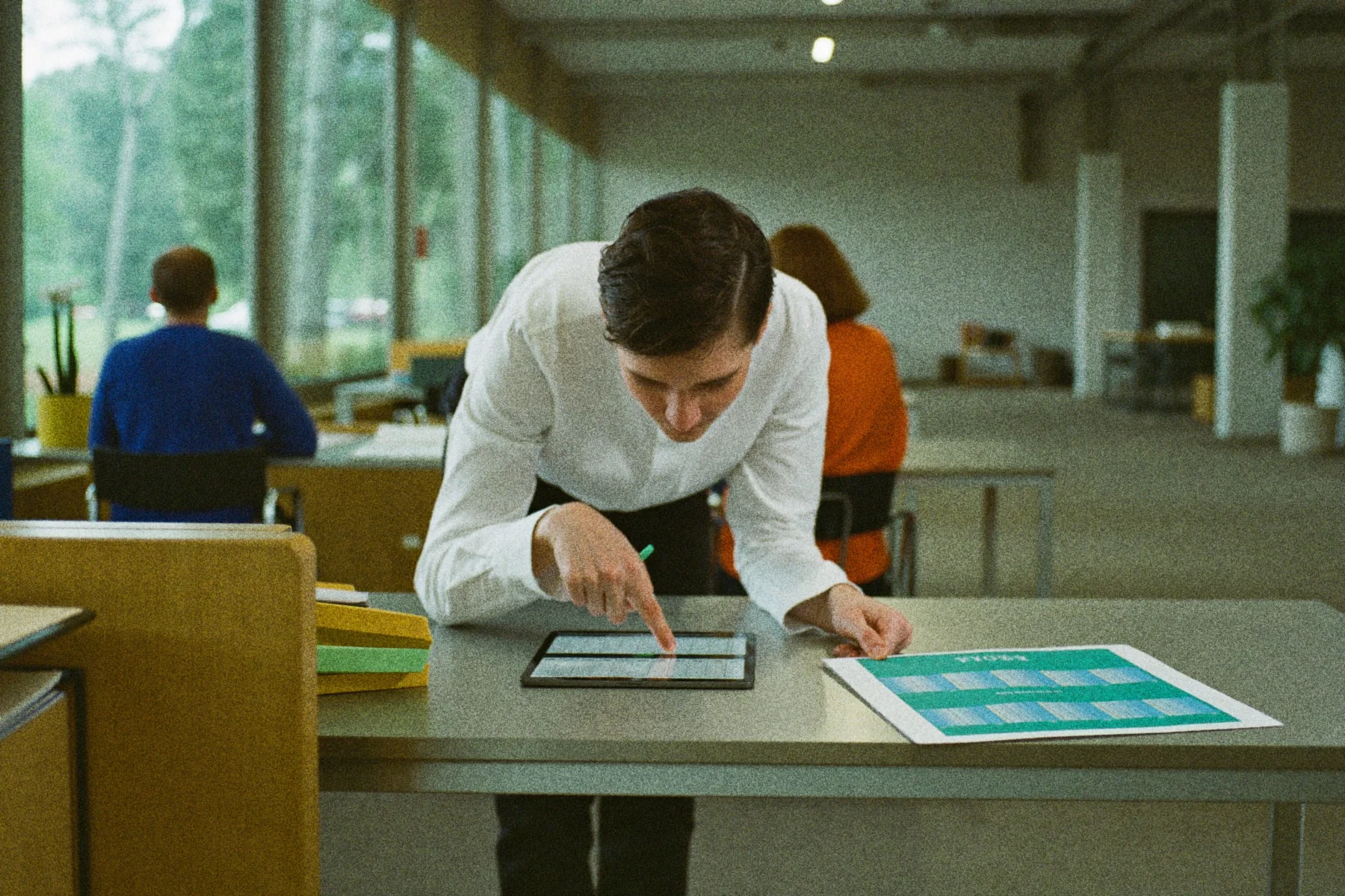 Een planner wijst naar een rij op een tablet terwijl een papieren weekplanning naast hem op het bureau ligt.