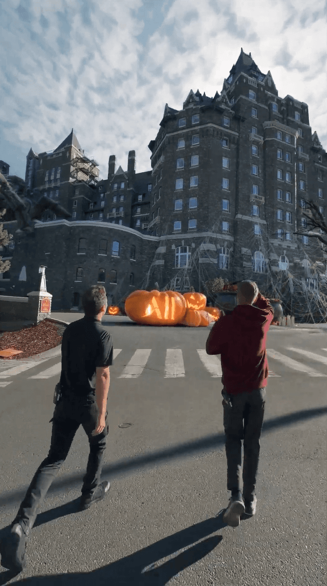 Two men looking at giant, glowing jack-o'-lanterns in front of a large stone building.