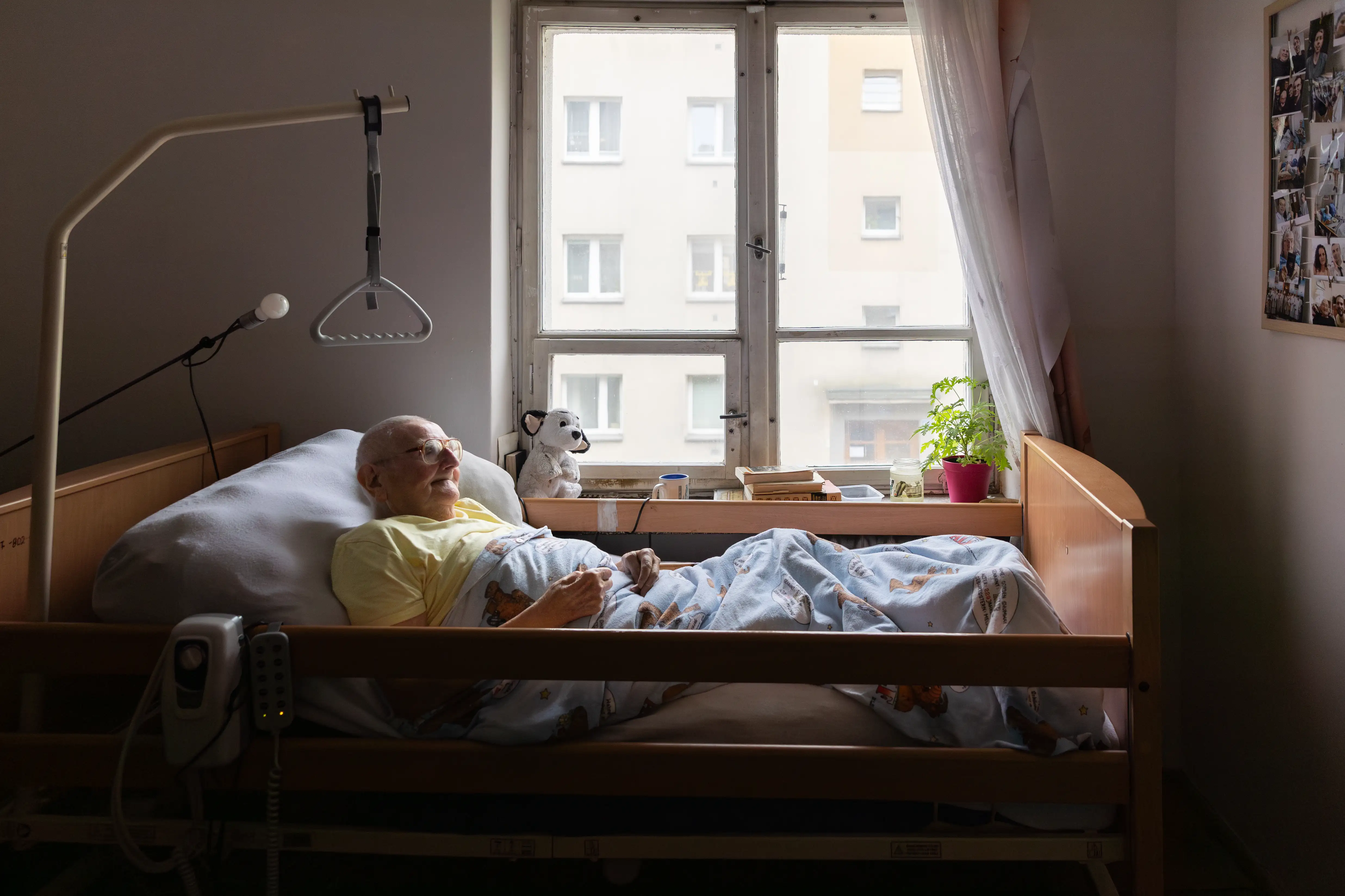 Starsza, niepełnosprawna osoba leżąca w łóżku, z dźwigiem do pomocy. Za oknem widnieją blokowiska.