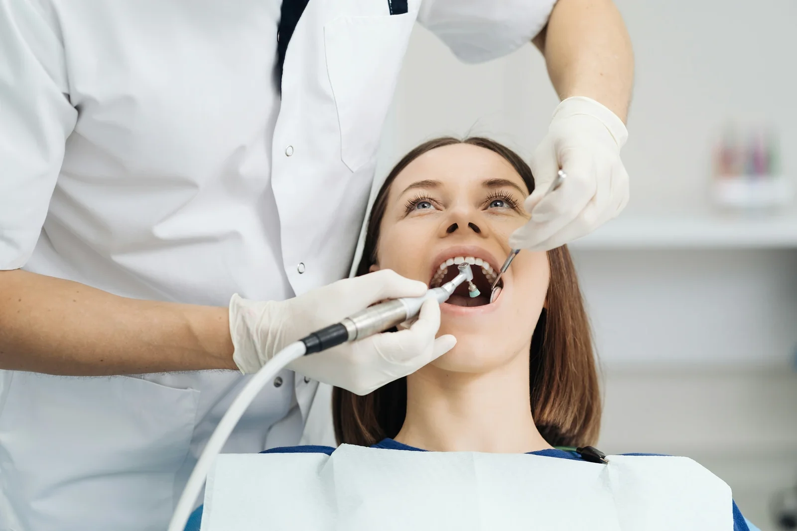 Dentist in white coat and gloves examines patient's teeth with dental tools in clinical setting.